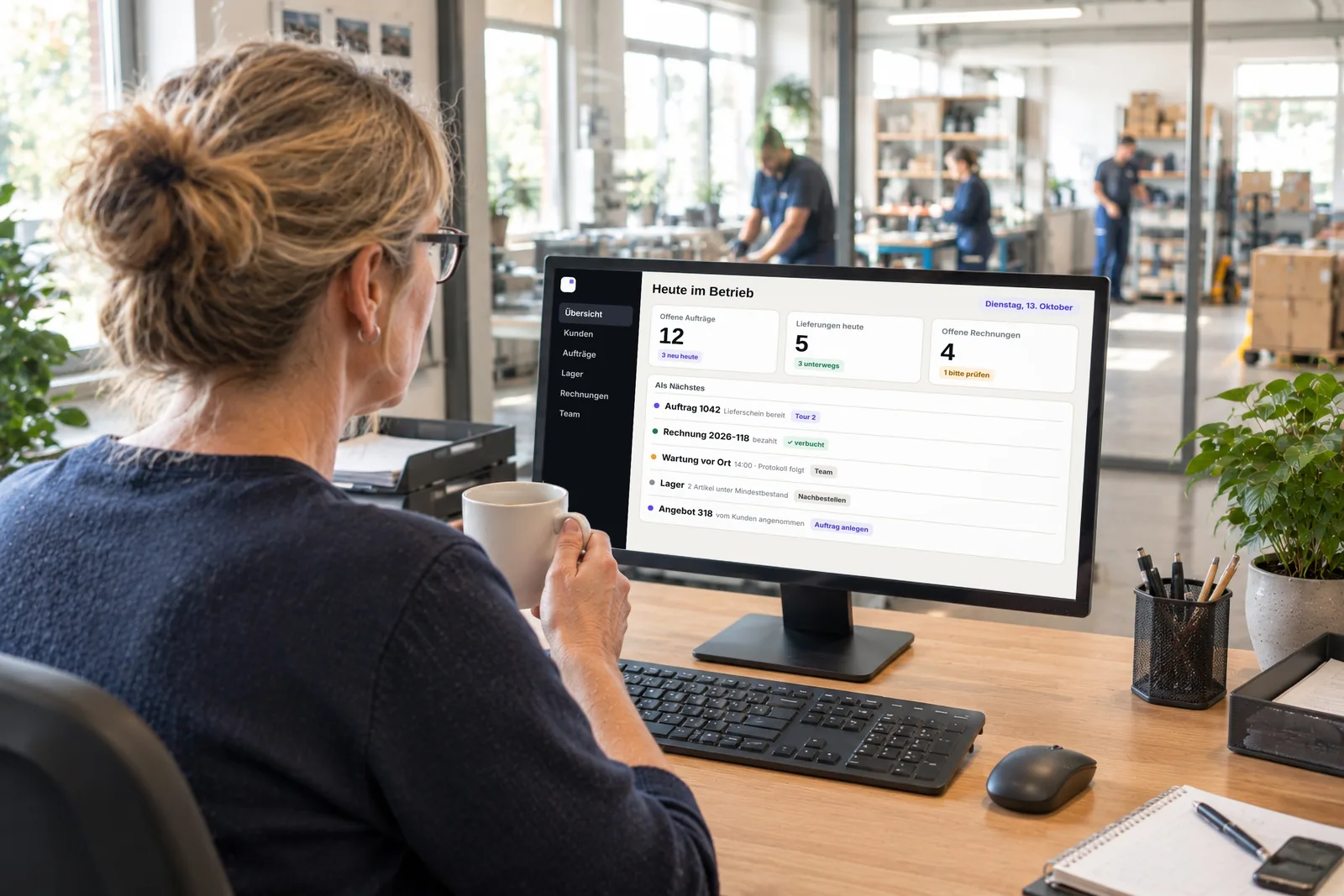 Eine Inhaberin sieht am Monitor die Tagesübersicht mit offenen Aufträgen, Lieferungen und Rechnungen. Hinter der Glaswand arbeitet ihr Team. Symbolbild, KI-erzeugt.